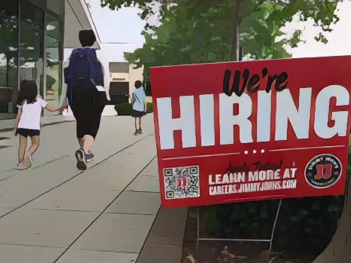 Menschen gehen an einem Schild mit der Aufschrift „Now Hiring“ vorbei, das vor einem Restaurant in Arlington, Virginia, angebracht ist.